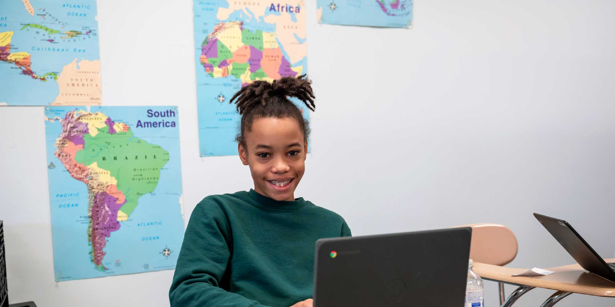 Smiling student working on a Chromebook in a classroom with world maps on the wall.